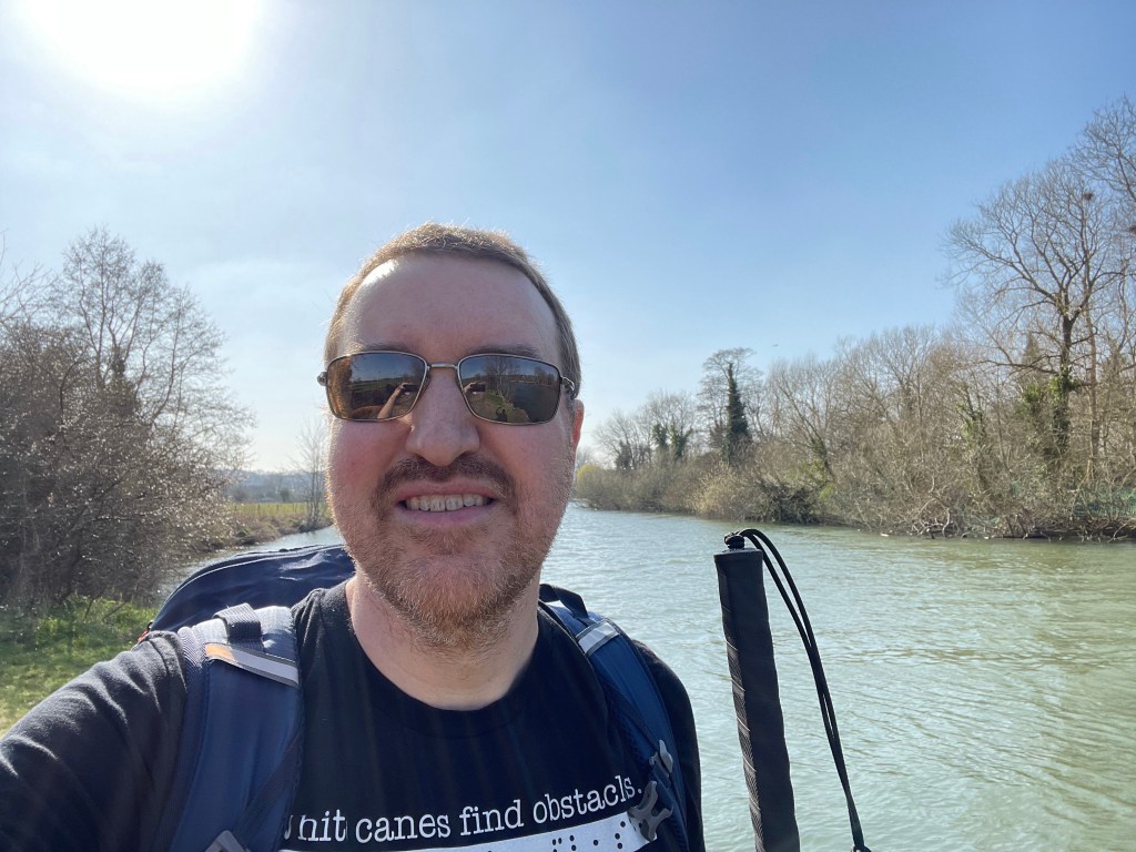 Chris, a white man with short brown hair and a greying beard is stood wearing dark sunglasses and holding a long white cane in front of a river under a clear blue sky