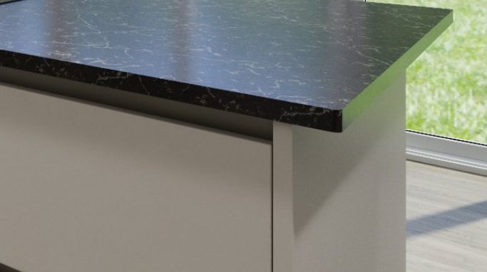 A closeup of a pebble-grey kitchen cabinet beneath a marbled black worktop. There is a groove beneath the worktop and a cut-away section across the top of the cabinet for a hand hold