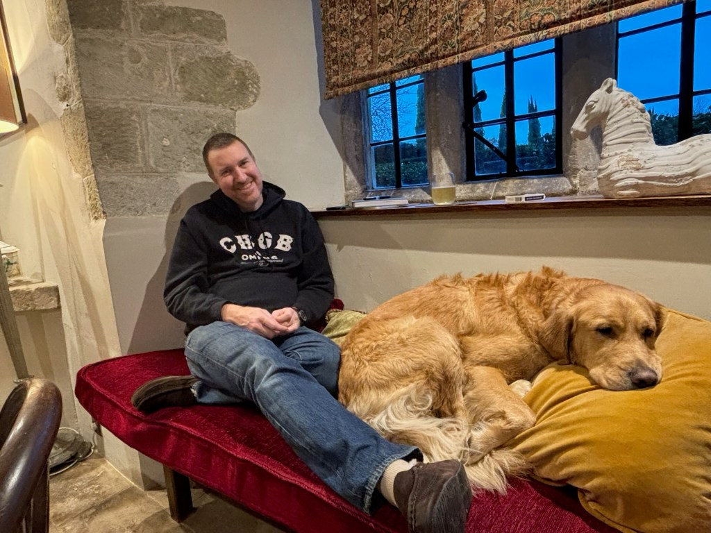 Chris and Chester sat on a long alcove beneath leaded windows in a 13th Century country house. Chris is rested back against the wall with his feet up and Chester is curled up beside his feet with his head rested on a cushion